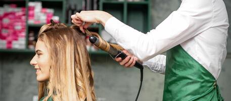 creëren krullen met curling strijkijzers. kapper maakt een kapsel voor een jong vrouw met lang rood haar- in een schoonheid salon. foto