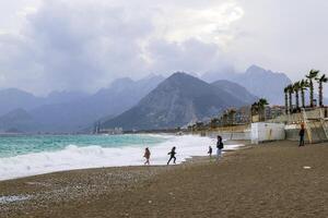 antalya, kalkoen - maart 05, 2023. onherkenbaar mensen zijn wandelen en spelen Aan dijk in de buurt naar stormachtig zee met groot golven en bergen Aan achtergrond. foto