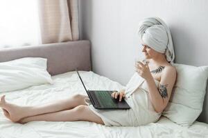 tver, rusland-augustus 2, 2021 een vrouw na een douche is aan het liegen Aan de bankstel met een laptop en een oog masker Bij de dezelfde tijd. de concept van multitasking in de eenentwintigste eeuw. foto