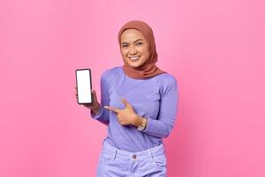 portret van glimlachende jonge aziatische vrouw die met de vinger op het scherm van een mobiele telefoon wijst op een roze achtergrond foto