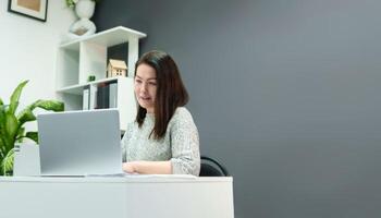 professioneel vrouw voert bedrijf correspondentie terwijl zittend Bij een bureau met een computer in een elegant kantoor met een grijs muur, kopiëren ruimte foto