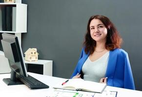 professioneel meisje. in een kantoor omgeving, een krullend haar brunette in een blauw pak werken professioneel Bij een computer, demonstreren haar kennis en vaardigheden foto