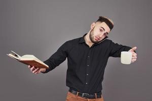 portret van jonge knappe man met telefoon, kopje koffie en boek op handen. het concept van overwerk, stress en veel werk foto