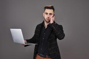 drukke bebaarde man in zakelijke kleding praten via smartphone en het gebruik van laptopcomputer terwijl hij een kopje koffie in de hand houdt over grijze achtergrond foto