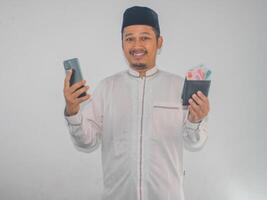 Aziatisch moslim Mens glimlachen gelukkig terwijl Holding mobiel telefoon en tonen papier geld van zijn portemonnee foto