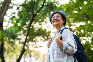 portret van Aziatisch volwassen vrouw in een hoed met rugzak achter haar rug, een Azië actief senior vrouw genieten van natuur in herfst park. staand Aan een spoor in een Woud buitenshuis. genieten van actief reizen reis foto