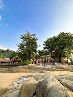 sereen tropisch strand tafereel met glad keien, gouden zand, en traditioneel hutten te midden van weelderig groen onder een Doorzichtig blauw lucht foto