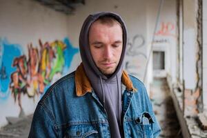 portret van jong knap vent in een denim jasje en een grijs kap poses tegen de achtergrond van een vernietigd en verlaten gebouw met graffitti. stedelijk concept. straat levensstijl. foto