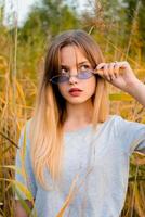 mooi jong meisje vervelend blanco grijs t-shirt en zwart jeans in blauw bril poseren tegen hoog groen en geel gras in vroeg warm herfst. buitenshuis portret van mooi vrouw model. foto