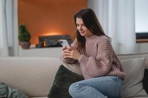 glimlachen vrouw op zoek Bij smartphone scherm, chatten online in sociaal netwerk met vrienden, zittend Aan comfortabel bankstel Bij huis, positief tevreden mooi jong vrouw surfing internet foto