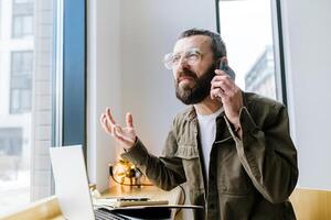 wtf. portret van een geschokt gebaard vent wie is pratend Aan een mobiel telefoon in een cafe. ver weg werk. freelancer foto