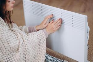 verkoudheid huis, bevriezen. gebruik makend van kachel Bij huis in winter. vrouw opwarming haar handen zittend door apparaat en vervelend warm kleren. verwarming seizoen. foto