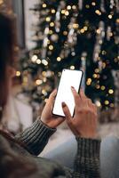 Kerstmis mockup met ruimte voor tekst. Kerstmis reclame, app sjabloon. handen Holding telefoon met leeg scherm Aan achtergrond van gouden mooi Kerstmis boom met lichten in avond foto