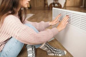 verkoudheid huis, bevriezen. gebruik makend van kachel Bij huis in winter. vrouw opwarming haar handen zittend door apparaat en vervelend warm kleren. verwarming seizoen. foto