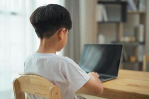 een jong jongen is zittend Bij een tafel met een laptop in voorkant van hem. hij is gefocust Aan zijn werk, mogelijk aan het doen huiswerk of aan het studeren. concept van concentratie en bepaling net zo de jongen werken Aan zijn taak foto