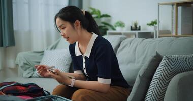 kant visie van Aziatisch tiener vrouw zittend Aan sofa maken checklist van dingen naar pak voor reis, voorbereiding reizen koffer Bij huis. foto