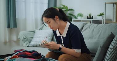 kant visie van Aziatisch tiener vrouw zittend Aan sofa maken checklist van dingen naar pak voor reis, voorbereiding reizen koffer Bij huis. foto