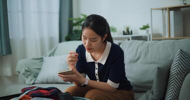 selectief focus van Aziatisch tiener vrouw zittend Aan sofa maken checklist van dingen naar pak voor reis, voorbereiding reizen koffer Bij huis. foto