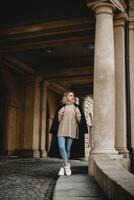 een modieus vrouw in een zwart jas staat vol vertrouwen in voorkant van een gebouw met een wit pijler. ze paren haar kleding met wit schoenen en blauw jeans, uitstralend stedelijk chique foto