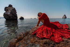 mooi sensueel vrouw in een vliegend rood jurk en lang haar, zittend Aan een rots bovenstaand de mooi zee in een groot baai. foto