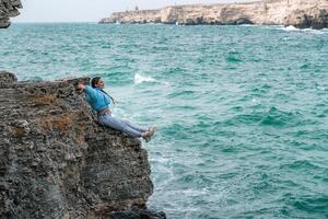 een vrouw in een blauw jasje zit Aan een rots bovenstaand een klif bovenstaand de zee, op zoek Bij de stormachtig oceaan. meisje reiziger rust, denkt na, dromen, geniet natuur. vrede en kalmte landschap, winderig het weer. foto