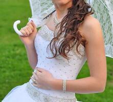 detailopname portret van jong mooi bruid in een bruiloft jurk staand Aan een groen veld- en Holding een wit paraplu foto
