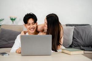 jong paar is sharing een vrolijk moment Aan een bankstel met een laptop. de Mens, gekleed in een wit shirt, lacht terwijl de vrouw, in een mouwloos bovenkant, leunt in met een aanhankelijk glimlach. foto