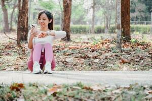 vrouw gezeten Aan een park pad looks Bij haar geschiktheid tracker terwijl Holding een smartphone, met verspreide herfst bladeren in de achtergrond. foto