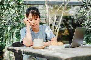 vrouw verschijnt benadrukt of attent terwijl werken Bij een buitenshuis houten tafel met een laptop en ontbijt, omringd door tuin groen. foto