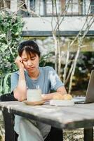 vrouw verschijnt benadrukt of attent terwijl werken Bij een buitenshuis houten tafel met een laptop en ontbijt, omringd door tuin groen. foto