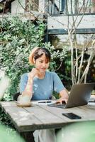 vrouw werken ijverig Aan haar laptop Bij een houten tuin tafel, genieten van een sereen buitenshuis atmosfeer. foto