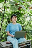 gelukkig vrouw zittend Aan een houten bank, werken Aan een laptop in een levendig tuin, genieten van afgelegen werk. foto