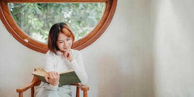 introspectief jong vrouw verdiept in lezing een boek, gezeten comfortabel door een circulaire venster met natuurlijk licht streaming in. foto