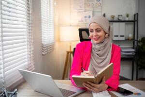 bedrijf Aziatisch moslim vrouw gebruik makend van rekenmachine en schrijven maken Notitie met berekenen financiën Aan een kantoor bureau. vrouw werken Bij kantoor met laptop en belasting, boekhouding, documenten Aan bureau foto