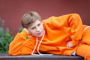 een tiener- jongen in oranje kleren poses terwijl aan het liegen Aan een bank. foto