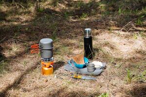 buitenshuis camping voedsel in zonnig het weer, toerist gebruiksvoorwerpen, een pot met een deksel, thee in een thermosfles, een mok staand Aan de grond, een geel mes, een gas- brander, een rust uit in de Woud. foto