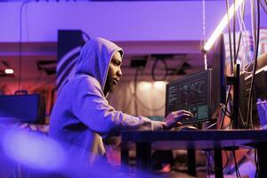 Afrikaanse Amerikaans hacker stelen gevoelig informatie van server Aan computer. Mens in kap ontwikkelen kwaadaardig software en overtreden gegevens in verlaten magazijn schuilplaats foto