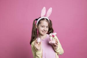 speels jong meisje poseren met gekleurde eieren en een gevuld roze konijn, vervelend konijn oren en glimlachen in voorkant van camera. klein aanbiddelijk kind tonen haar feestelijk Pasen decoraties en speelgoed. foto