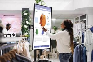 vrouw koper selecteren kleren Aan bord, gebruik makend van interactief tintje scherm toezicht houden op in kleding op te slaan. jong winkel klant buying mode items van zelf bestellen kiosk onderhoud, kleinhandel winkel. foto