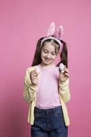 klein kind poseren met vertrouwen in voorkant van camera, tonen haar geschilderd kleurrijk eieren voor Pasen viering. jong zoet kind met konijn oren glimlachen in studio, presenteren handgemaakt decoraties. foto