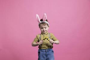 blij klein kind spelen met een kleurrijk speelgoed- in voorkant van camera, genieten van vrije tijd werkzaamheid tegen roze achtergrond. jong vrolijk jongen vervelend konijn oren en poseren voor een Pasen fotoshoot. foto