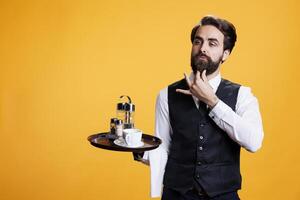 elegant butler met pak en stropdas in studio, acteren zelfverzekerd terwijl hij houdt voedsel schotel met bestek en koffie kop naar dienen klanten. professioneel geschoold ober poseren Aan camera, gevoel toegewijd. foto