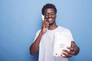 jong zwart Mens staand tegen geïsoleerd blauw achtergrond is beantwoorden telefoon telefoongesprek. portret van glimlachen Afrikaanse Amerikaans vent genieten van een vaste telefoon telefoon gesprek met geliefde degenen. foto