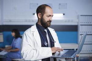 Kaukasisch dokter is staand en zoeken voor iets Aan zijn persoonlijk laptop, in medisch kantoor. in achtergrond, verpleegster gebruik makend van bureaublad pc met gedetailleerd focus Aan mannetje arts Holding een minicomputer. foto