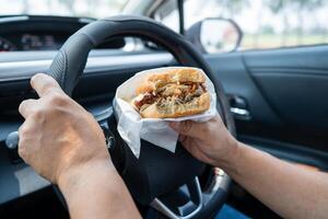 Aziatisch dame Holding Hamburger en Frans Patat naar eten in auto, gevaarlijk en risico een ongeluk. foto