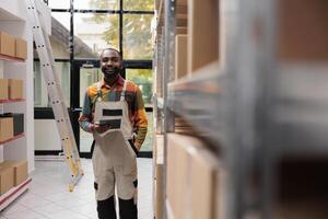 glimlachen supervisor staand in de buurt karton dozen, werken Bij producten voorraad in Magazijn. pakhuis arbeider vervelend industrieel algemeen terwijl voorbereidingen treffen klanten bestellingen voordat levering foto