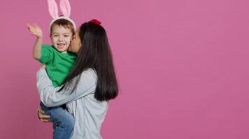 moeder hijs- omhoog haar weinig jongen en zoenen hem in studio, tonen liefde en knuffelen schattig kind tegen roze achtergrond. zoet mam en zoon omarmen elk andere en lachend. camera a. foto