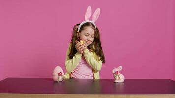 blij weinig meisje spelen met feestelijk Pasen decoraties in studio, creëren regelingen met een kuiken, konijn en ei. glimlachen schattig kleuter met konijn oren tonen kleurrijk ornamenten. camera b. foto