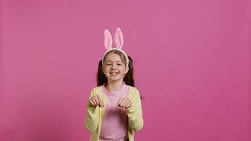 blij zorgeloos schoolmeisje jumping in de omgeving van in studio, imiteren een konijn en hoppen tegen roze achtergrond. vrolijk actief kind vervelend konijn oren en stuiteren, aanbiddelijk kind. camera b. foto
