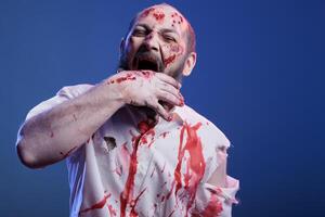 portret van duivels maniac zombie met bloed Aan gezicht en kleren bijten zijn hand, gevoel hongerig voor vlees. gek ondood monster kauwen Aan eigen arm, geïsoleerd over- studio achtergrond foto